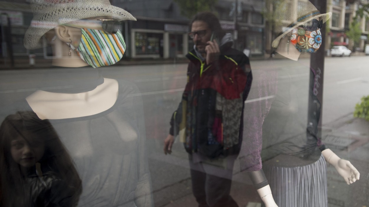 Un homme parle au téléphone cellulaire devant la vitrine d'un magasin avec deux mannequins qui portent un masque.