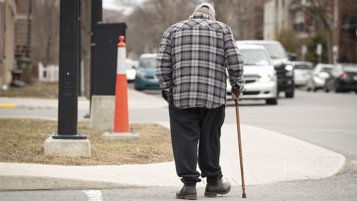Un homme âgé marche sur le trottoir avec une canne, dans une rue où passent des voitures.