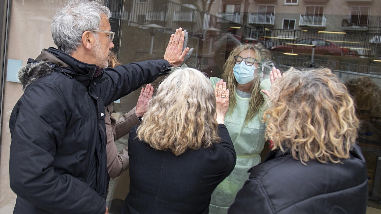 Une femme portant une jaquette jaune et un masque de protection regarde un homme et quatre femmes par la fenêtre.