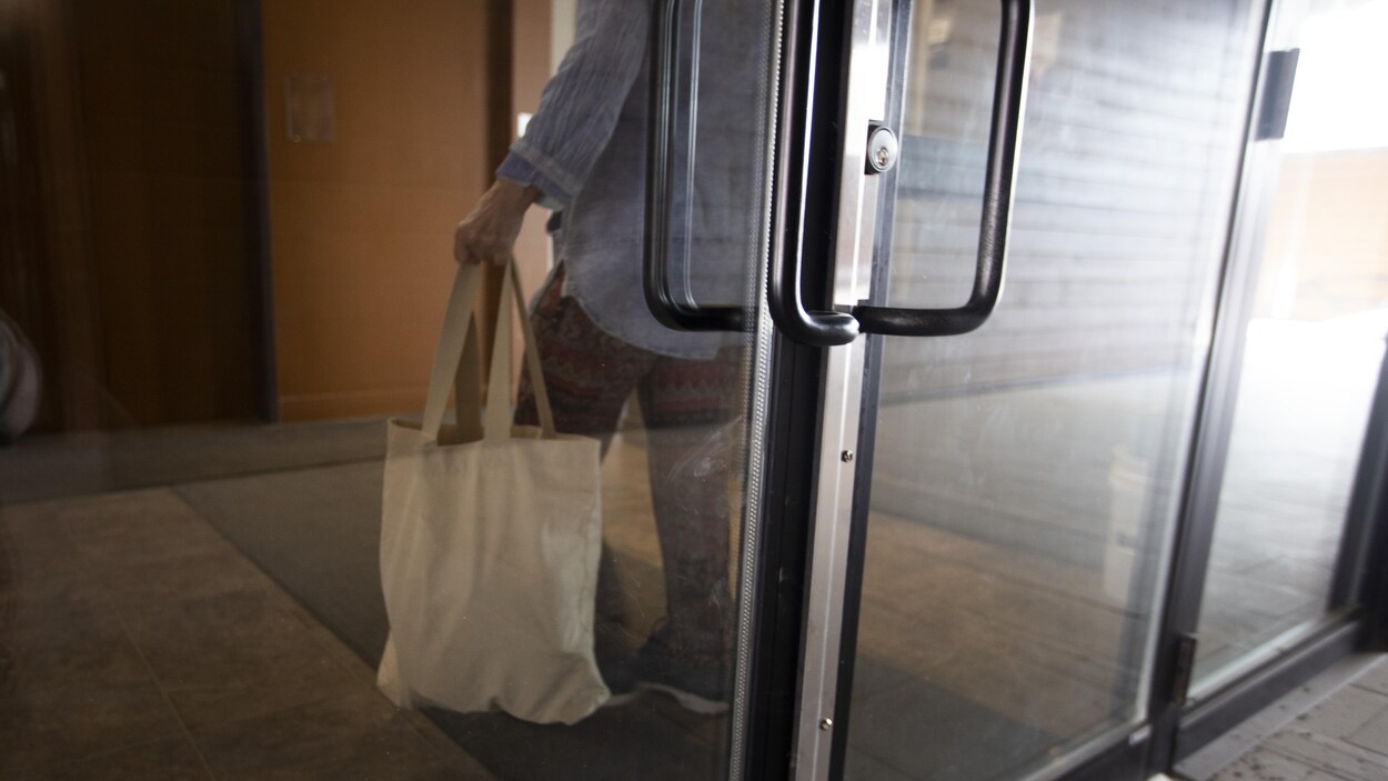 Une femme âgée transporte un sac de provision dans le hall d'entrée d'un immeuble, devant un ascenseur.