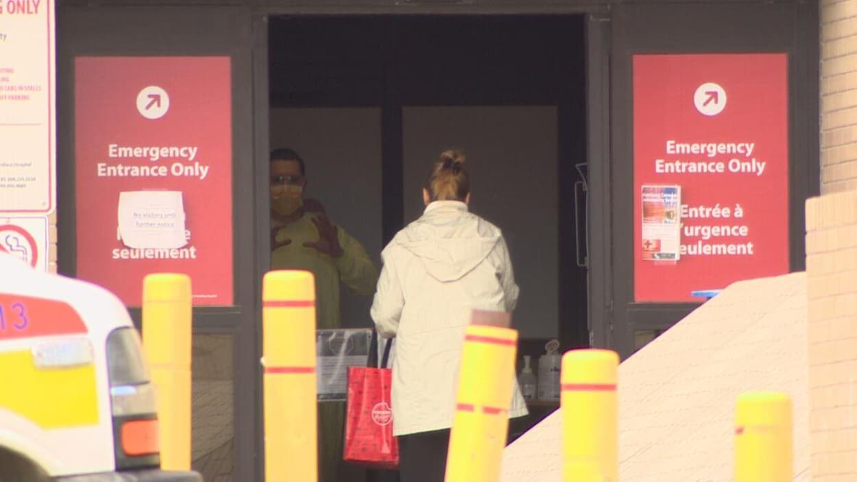 Une femme vue de dos est à l'entrée de l'urgence où un homme revêtu d'un équipement de protection l'accueille.