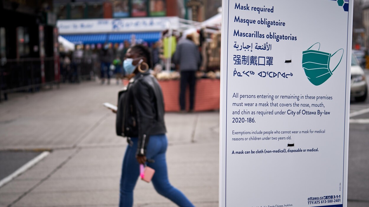Une femme marche dans la rue avec un masque. En premier plan, une pancarte d'avis publique de la Ville d'Ottawa.