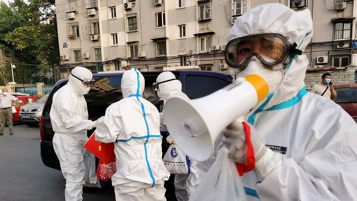 Du personnel en combinaison de protection dans la rue à Pékin, une personne parle dans un porte-voix.