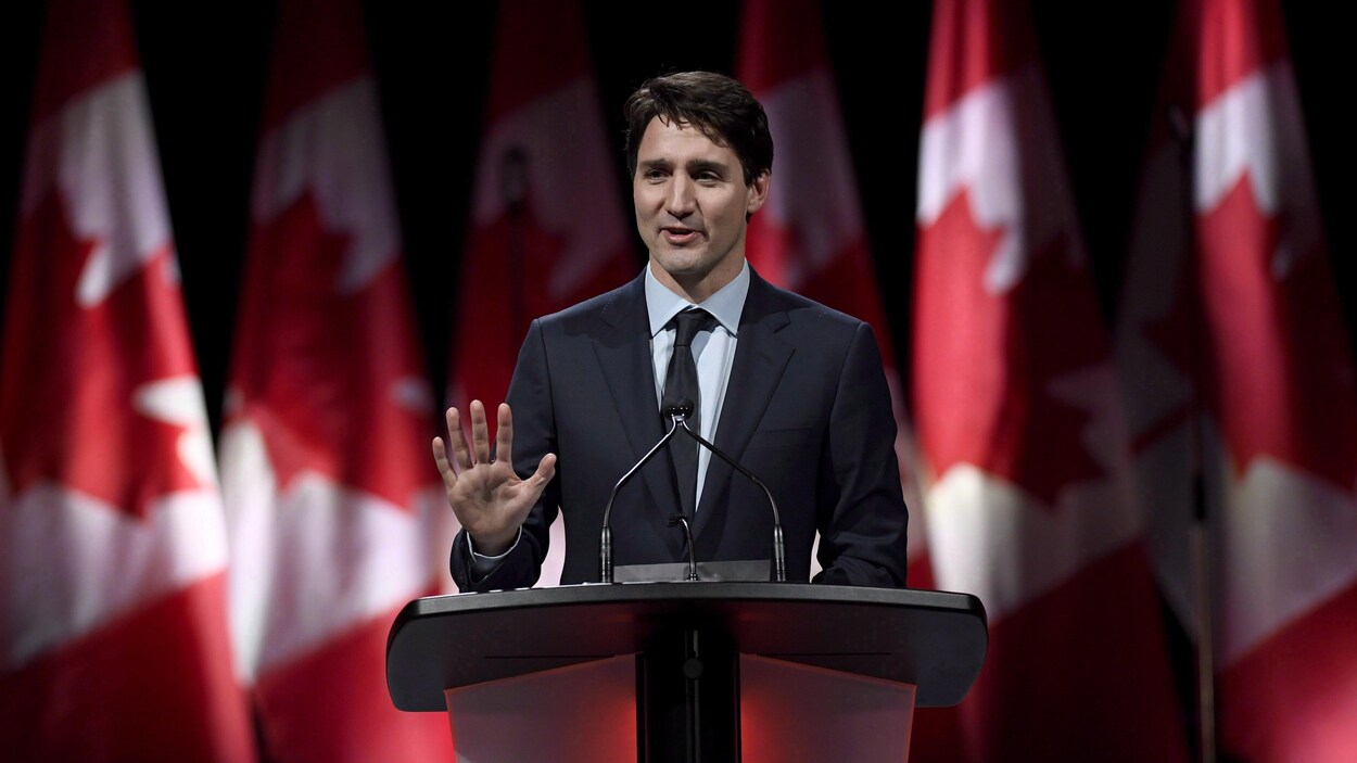Justin Trudeau sourit, levant la main, devant une série de drapeaux du Canada. Justin Trudeau sourit, levant la main, devant une série de drapeaux du Canada.