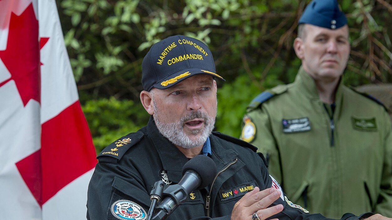 Un homme portant la casquette et un manteau de la marine canadienne parle au micro, sous le regard d'un autre homme en uniforme.