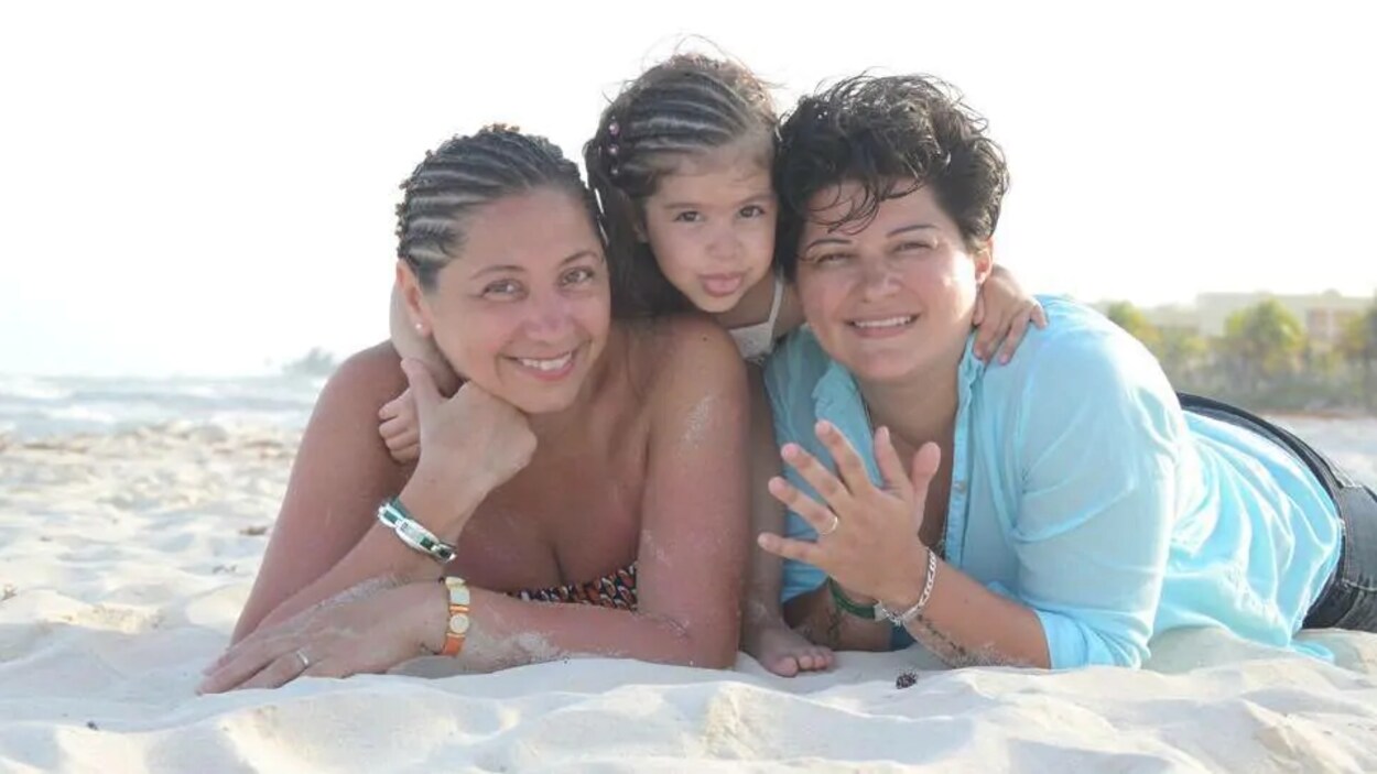 Deux femmes souriantes et une petite fille qui grimace étendues sur une plage de sable blanc.