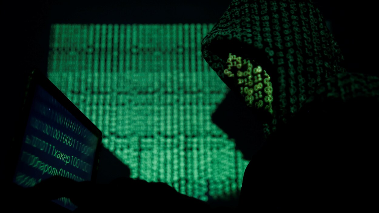 Un homme portant un capuchon, dont le visage est remplacé par du code informatique, est assis devant un écran où l'on voit aussi du code informatique. Un homme portant un capuchon, dont le visage est remplacé par du code informatique, est assis devant un écran où l'on voit aussi du code informatique.