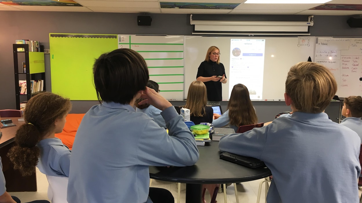 Une enseignante, avec un cellulaire entre les mains, se trouve face à une classe de sixième année. Des jeunes écoutent attentivement les explications de leur enseignante sur les risques des réseaux sociaux.