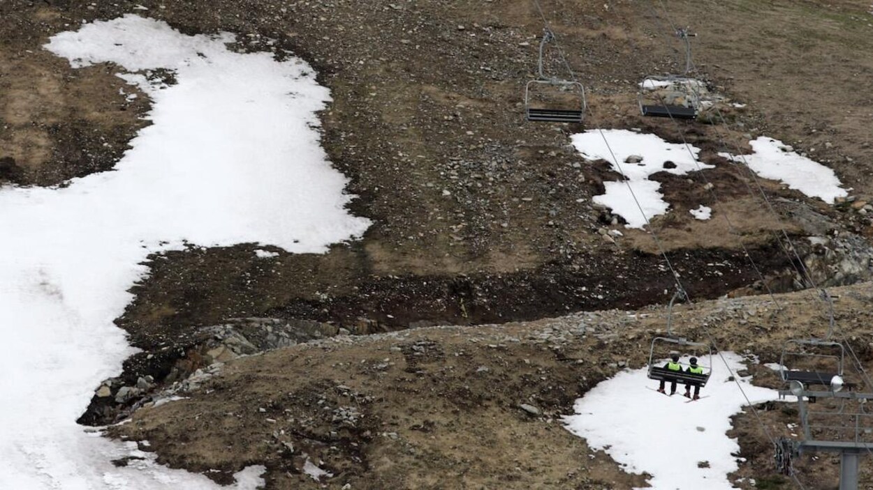 Un remonte-pente transporte deux skieurs au-dessus d'une montagne sur laquelle il n'y a pratiquement plus de neige et seulement de l'herbe et des roches.
