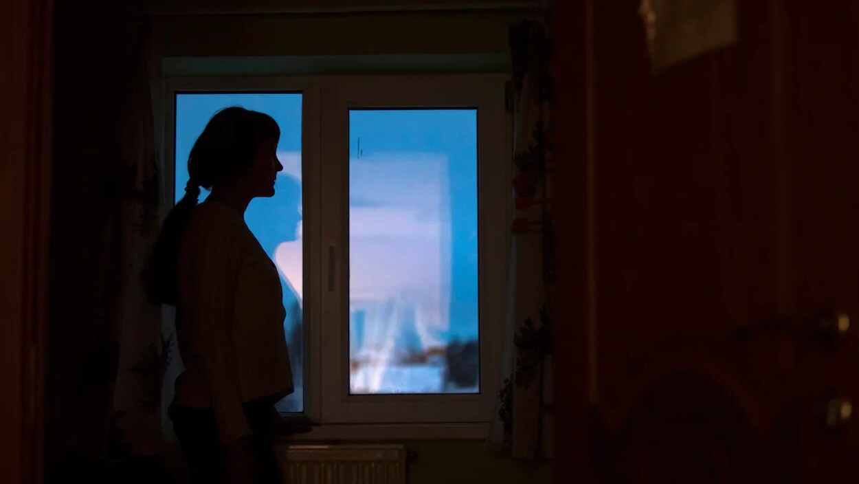 Vue sur une dame qui est debout à côté d'une fenêtre. Elle est dans le noir.