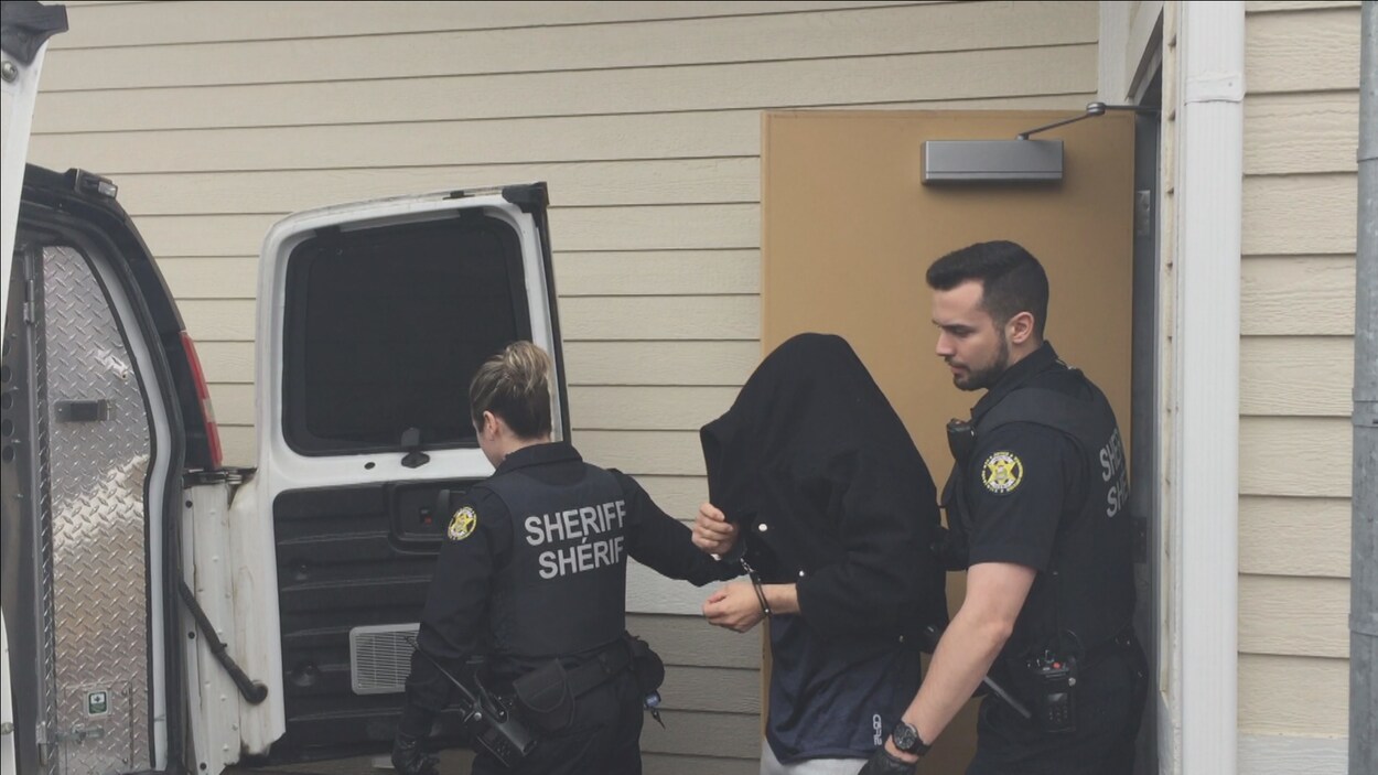 David Chiasson, un manteau cachant son visage, accompagné de deux policiers.