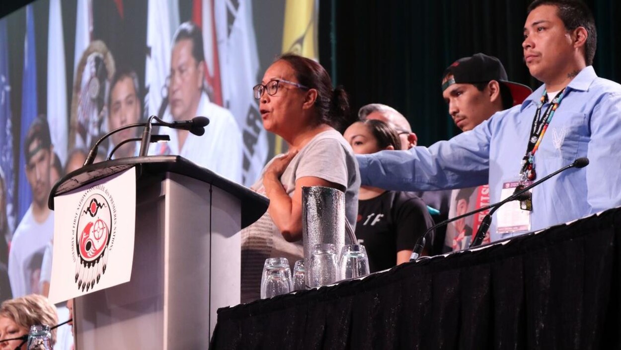 La mère de Colten Boushie fait un discours. Sa main gauche est posée sur son coeur, son visage est ému. Des gens se tiennent derrière elle, dont un homme qui pose une main dans le dos de la mère en signe de soutien.