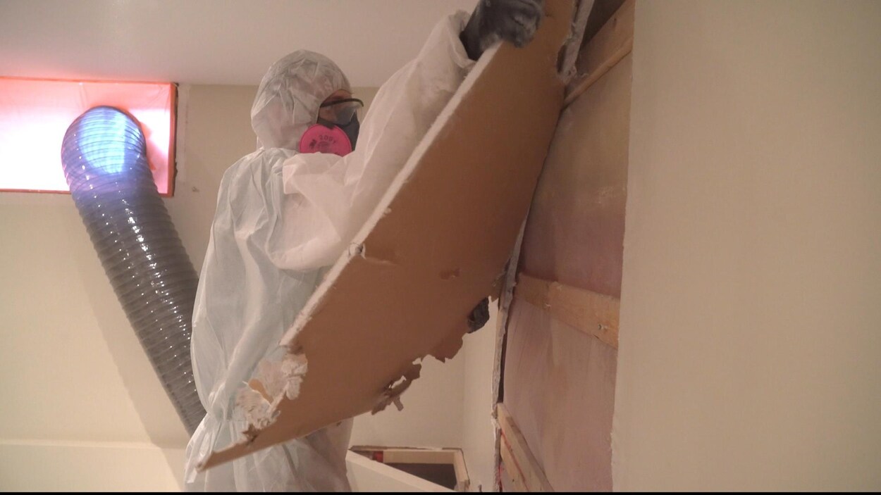 Un employé en habit de protection retire des panneaux de gypse d'un mur.