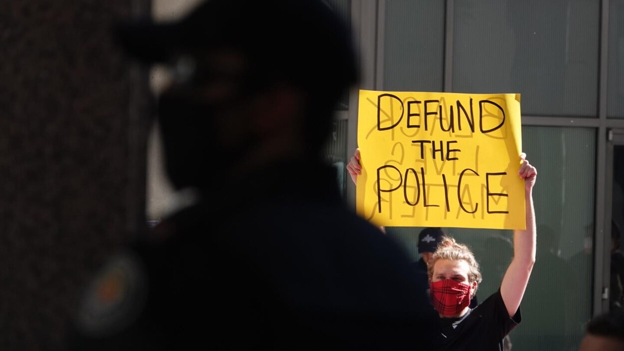 Un homme tient une pancarte sur laquelle est écrit « Defund the police » devant un policier.