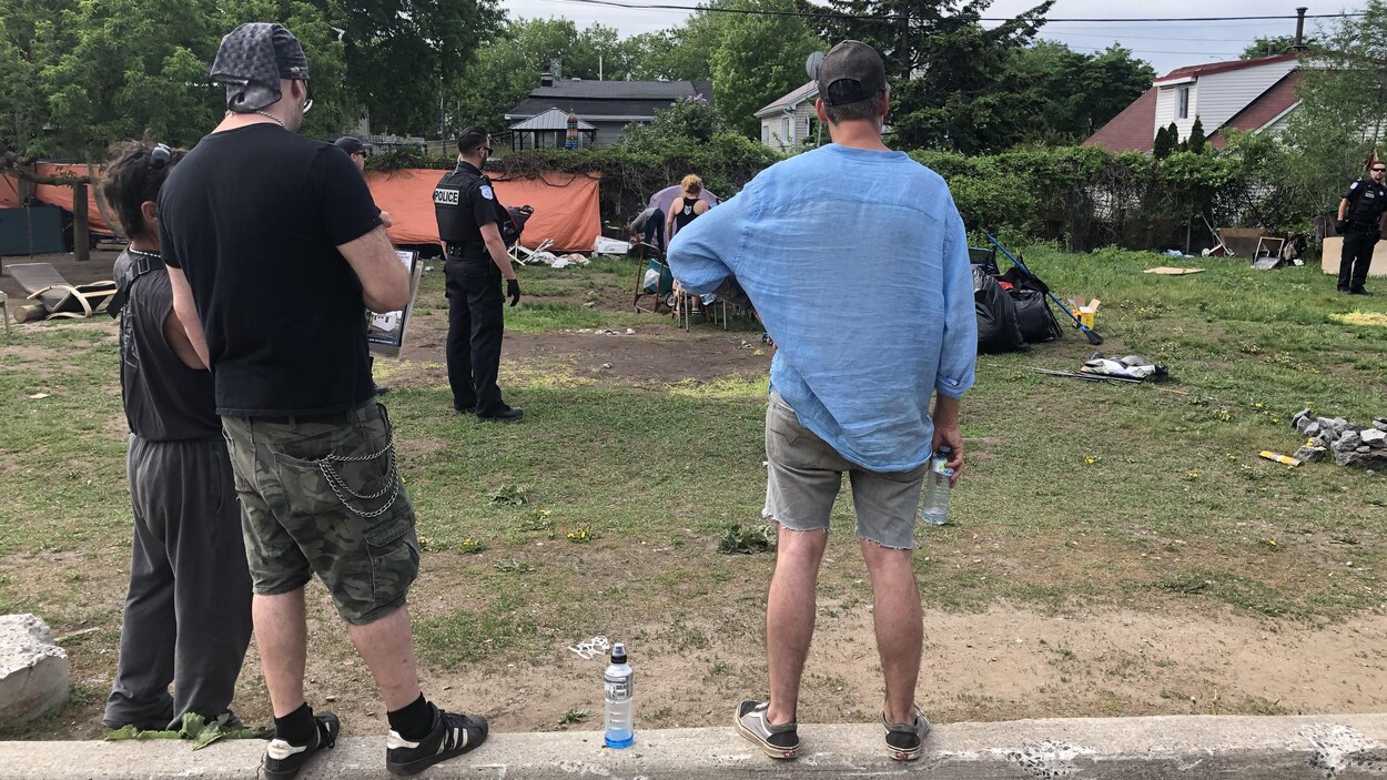 Trois hommes regardent des policiers démanteler le campement installé sur un terrain gazonné.