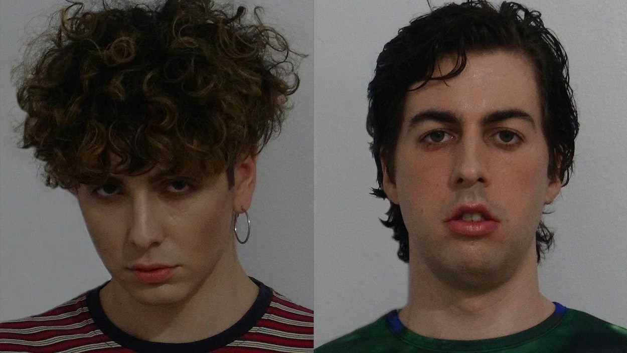 Les deux artistes fixent la caméra et se tiennent devant un fond gris.