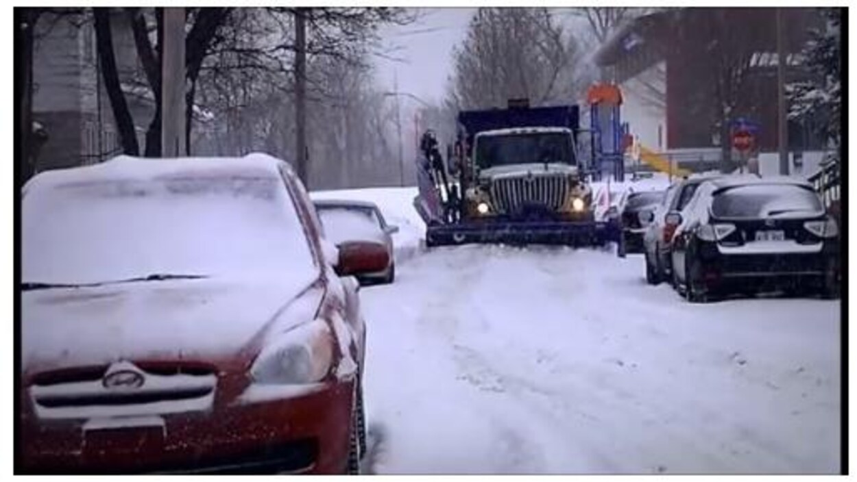 Une déneigeuse tente de se frayer un chemin dans une rue de la ville entre des voitures stationnées