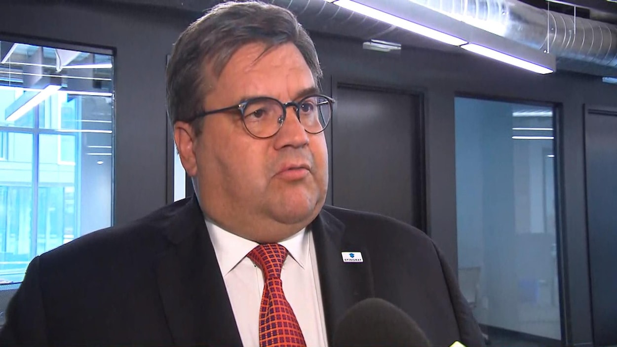 Un homme, qui semble costaud ou enrobé et qui porte des lunettes, regarde droit devant lui. Il semble parler à quelqu'un.