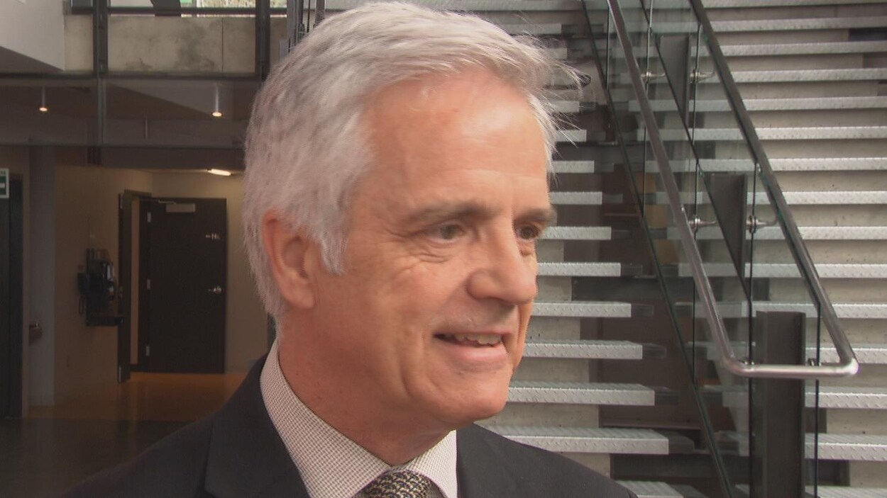 Le recteur parle aux journalistes dans un hall devant des escaliers.
