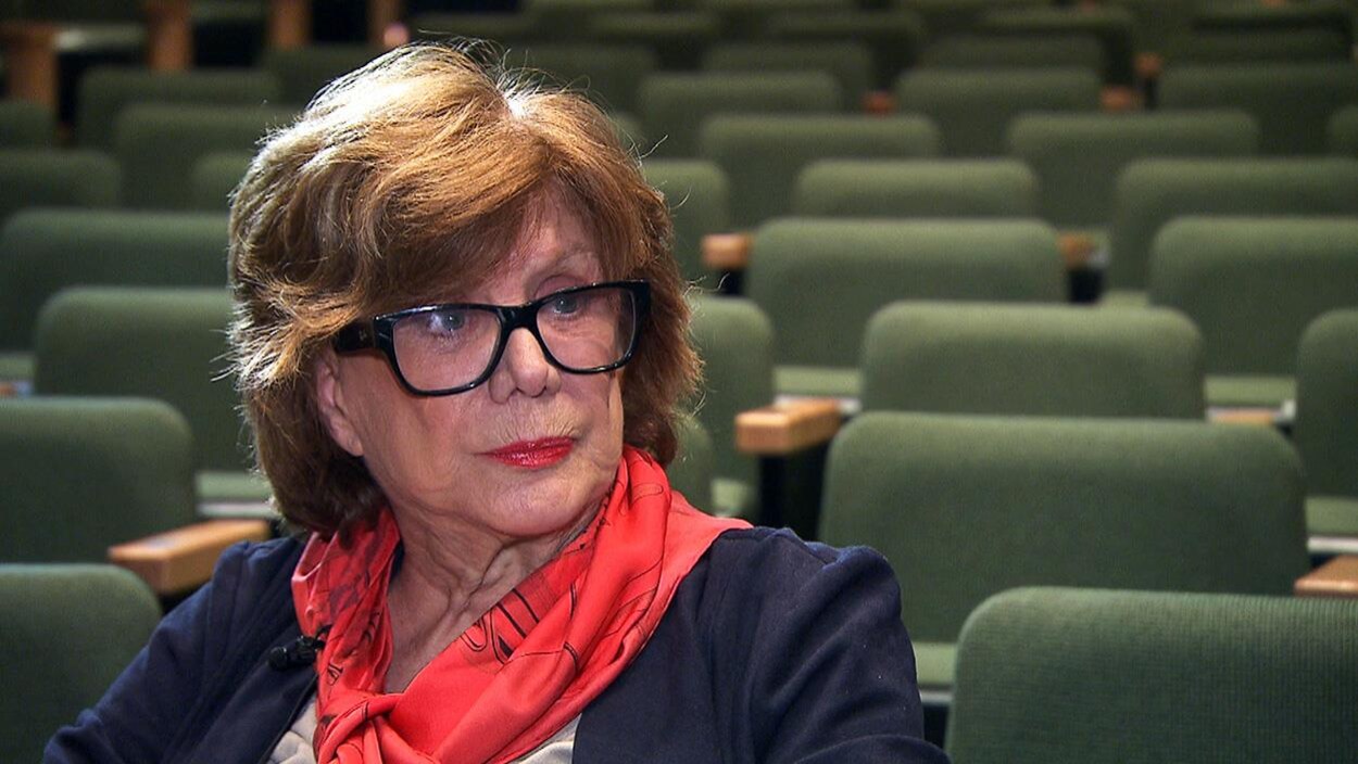 Denise Filiatrault porte des lunettes et est assise sur un banc dans une salle de théâtre vide.