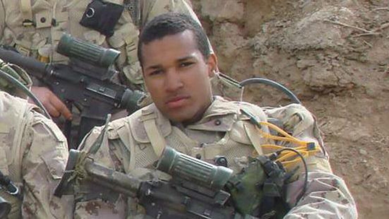 Lionel Desmond en uniforme militaire assis en Afghanistan, son fusil d'assaut repose sur ses genoux.