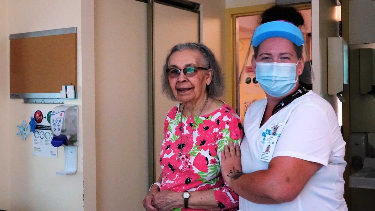 Une vieille dame au chemisier à fleurs rose, soutenue par une préposée avec un masque et une visière sur le visage.