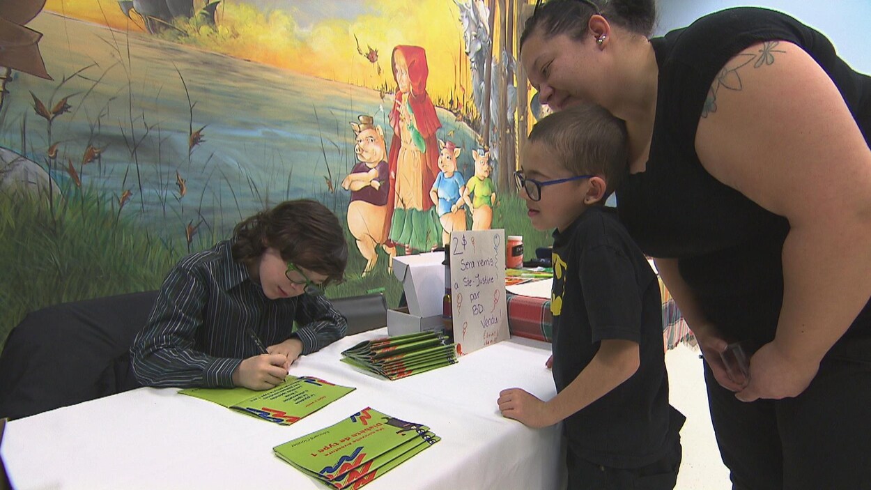Une jeune garçon derrière une table dédicace une bande dessinée à un autre jeune garçon.