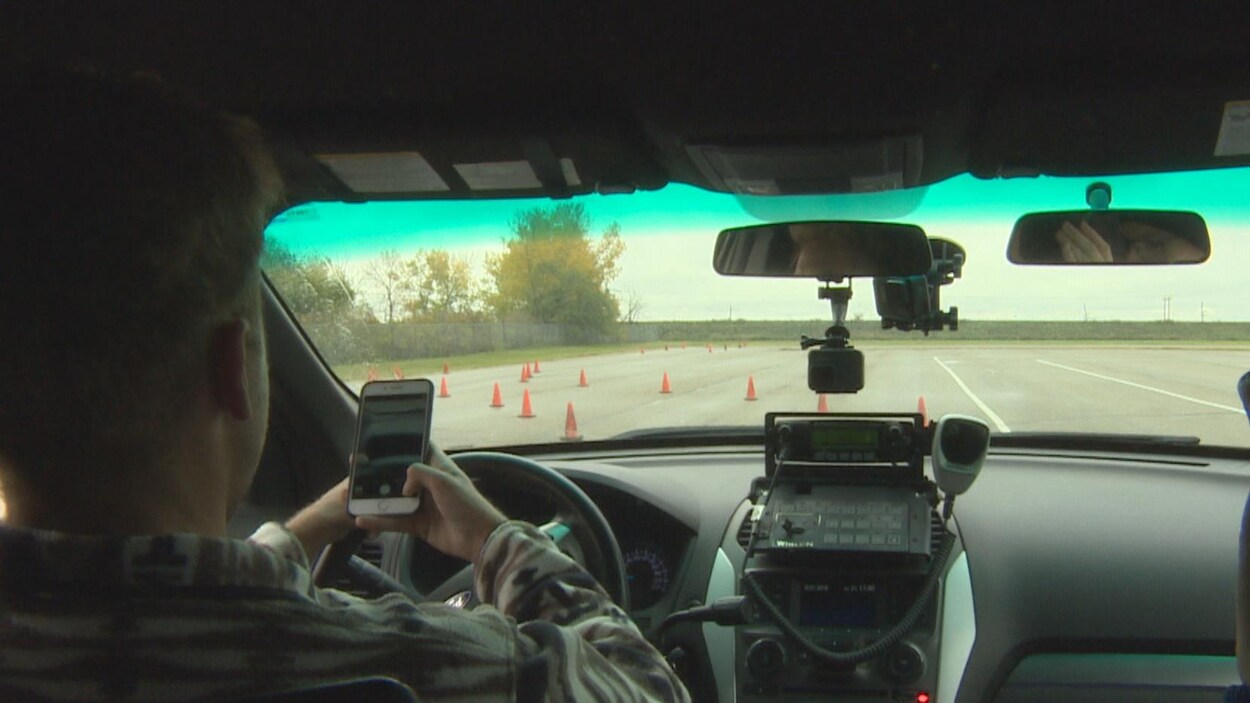 Un homme conduit avec un téléphone cellulaire à la main. Sur la route, il y a des cônes orange.