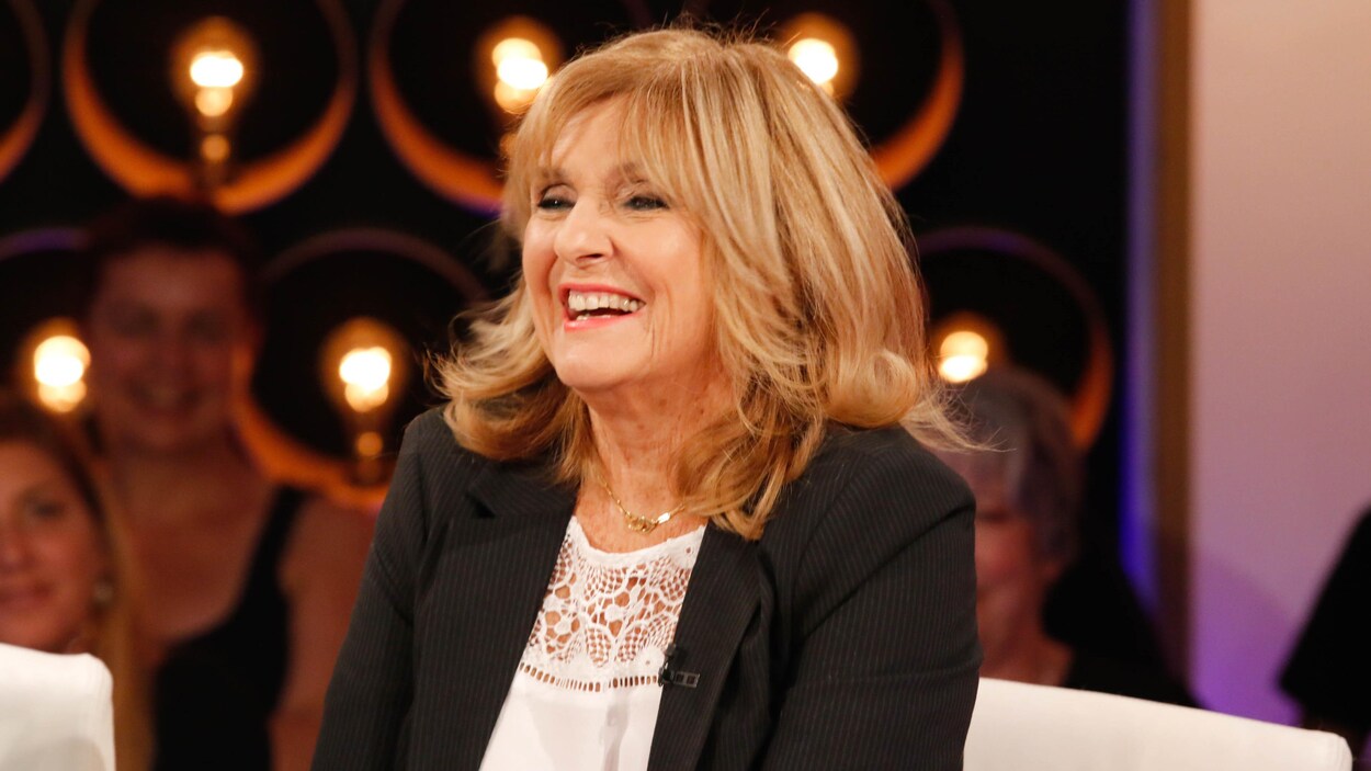 Dominique porte une blouse blanche et un veston noir à fines rayures.