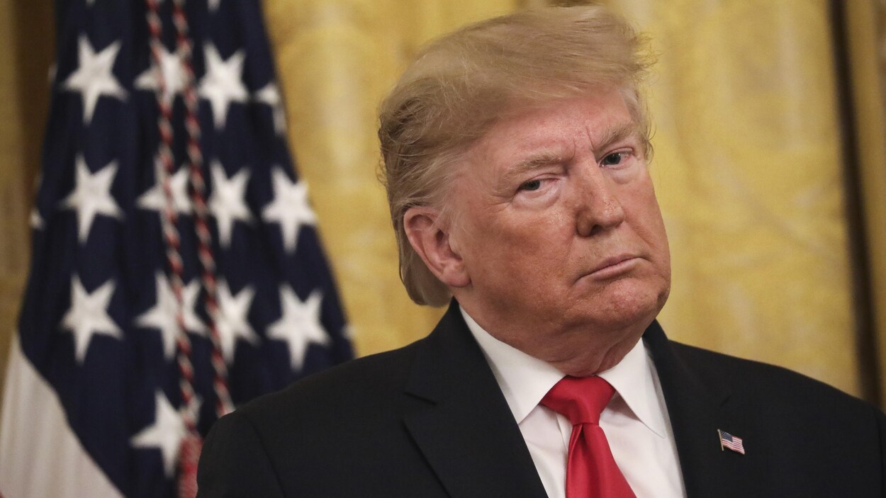 Le président des États-Unis Donald Trump se tient droit près d'un drapeau sur fond jaune.