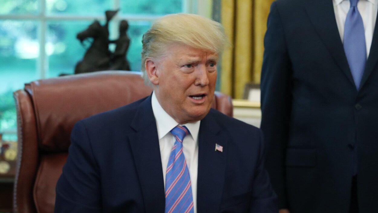 Le président Trump est assis derrière un bureau, les bras ouverts.