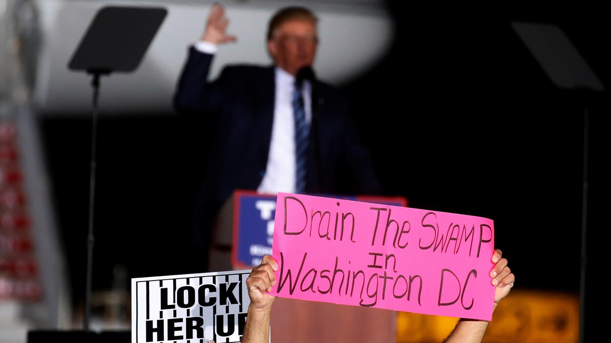 Une pancarte rose sur laquelle est écrit « Drain the swamp in Washington DC » est tenue devant Donald Trump, flou en arrière-plan. Une pancarte rose sur laquelle est écrit « Drain the swamp in Washington DC » est tenue devant Donald Trump, flou en arrière-plan.