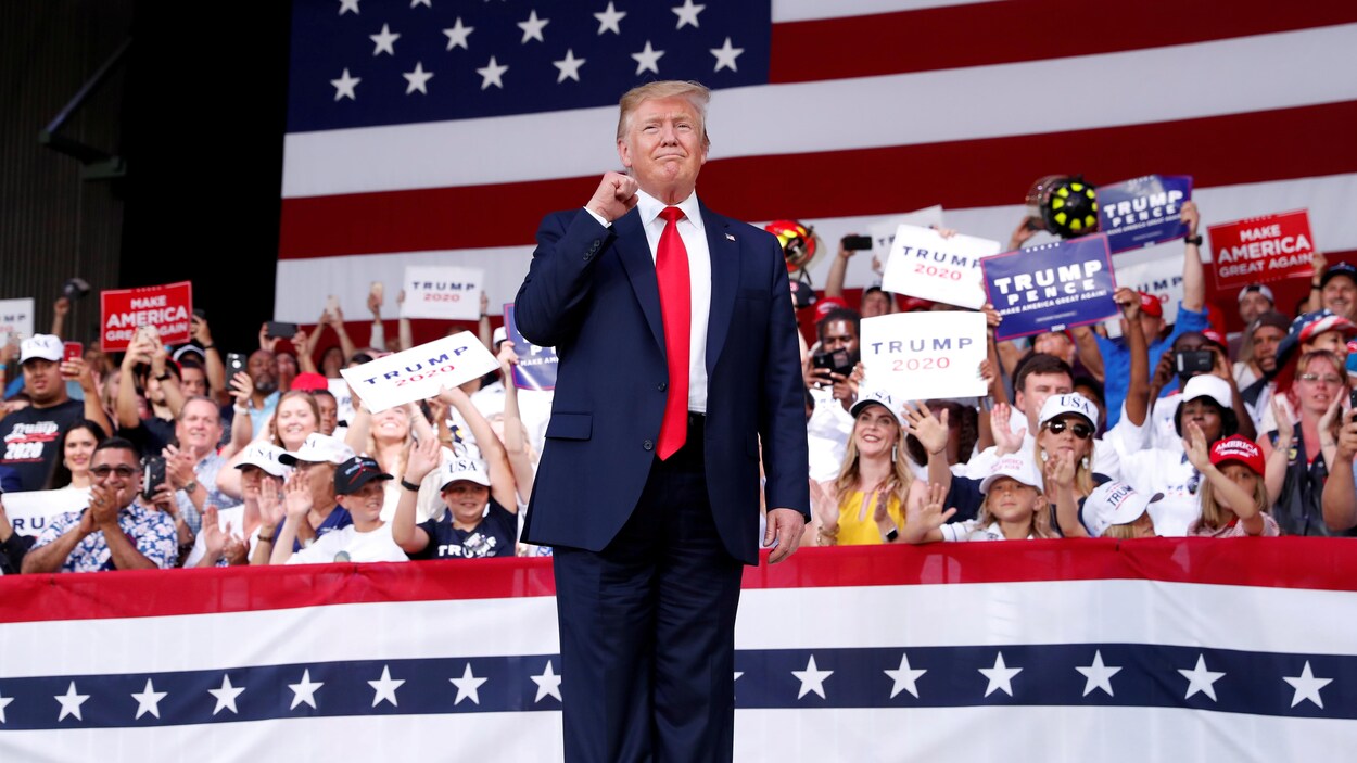 Donald Trump lève le poing sur une scène, entouré de partisans et du drapeau américain.