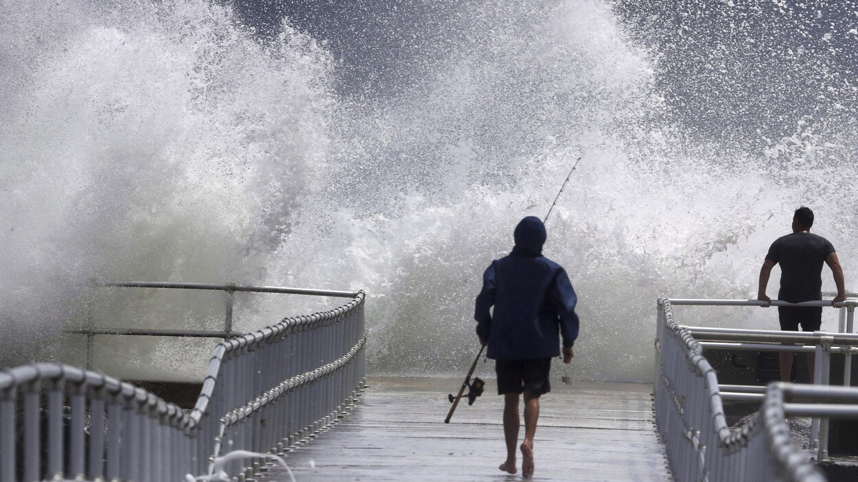 Des hommes marchent un quai où une vague gigantesque se brise dans les airs