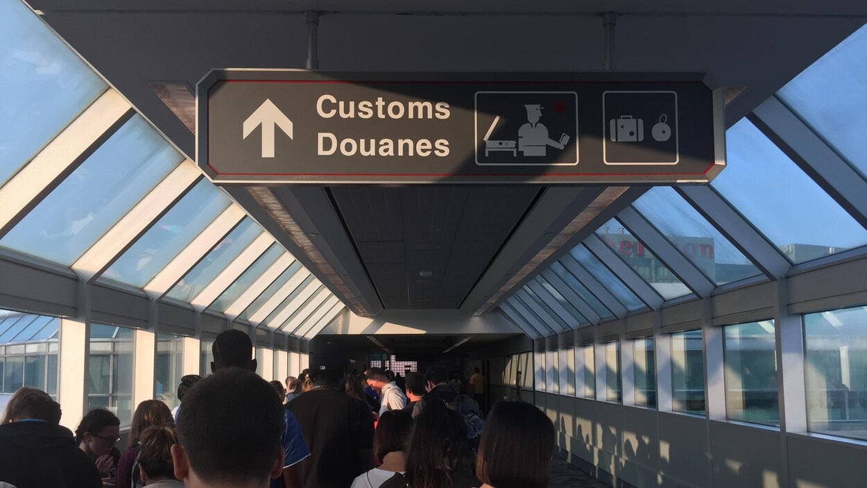 Plusieurs personnes attendent dans le corridor vitré menant aux douanes de l'aéroport international Pearson de Toronto; on voit une pancarte bilingue annonçant les douanes.