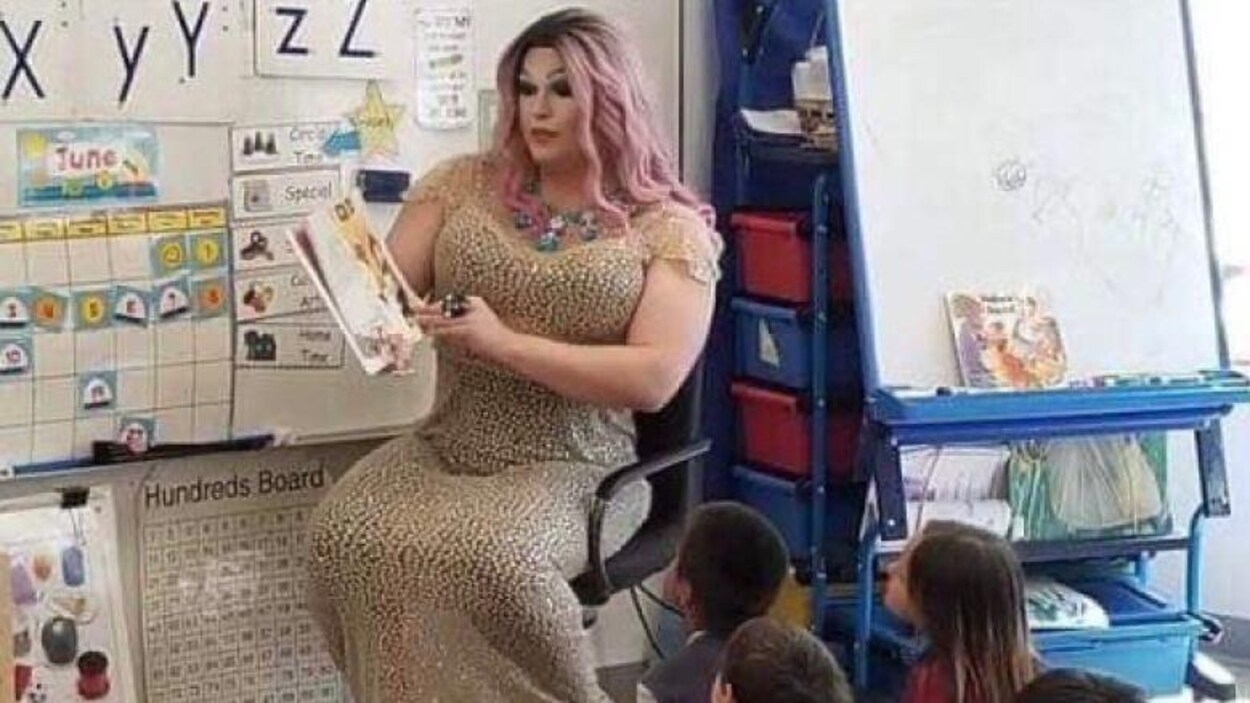 Jesse Ireland habillé en drag queen est assis sur une chaise dans une salle de classe faisant la lecture à des enfants assis à ses pieds.