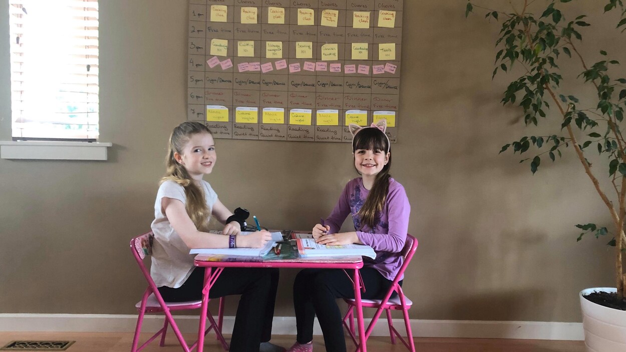 Deux filles sont assises à une petite table devant un grand tableau d'emploi du temps journalier.