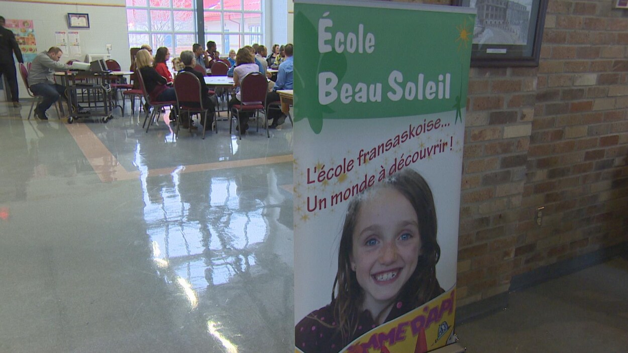 Une pancarte faisant la promotion de l'École Beau Seoleil, représentant le visage d'une petite fille souriante. En arrière plan, une salle avec des personnes assises à des tables. Une pancarte faisant la promotion de l'École Beau Seoleil, représentant le visage d'une petite fille souriante. En arrière plan, une salle avec des personnes assises à des tables.