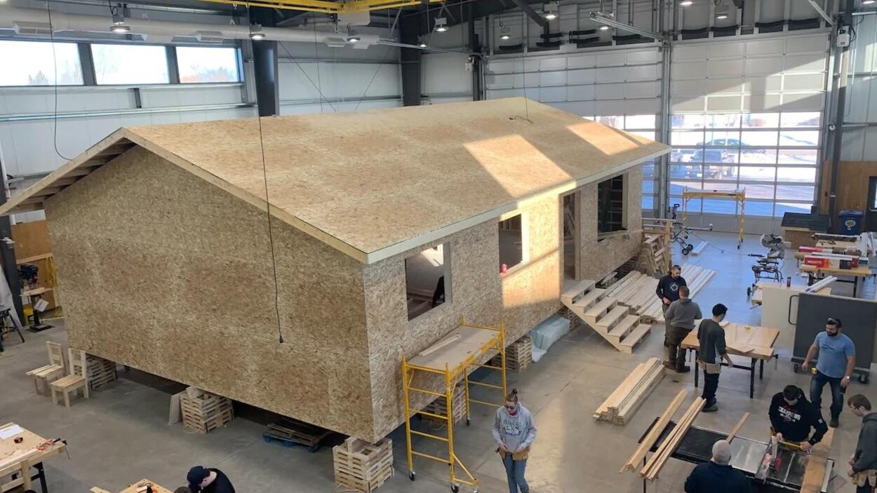 Une maison en construction est dans un vaste atelier, une douzaine de personnes travaillent autour.