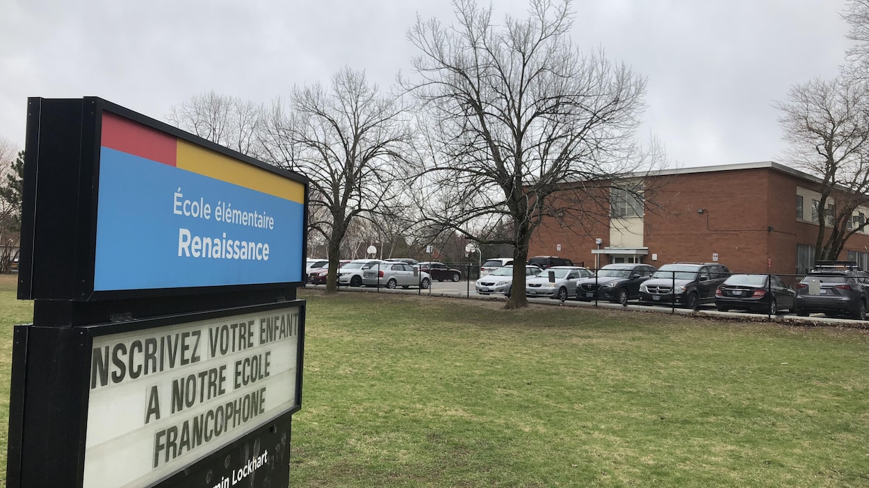 Une pancarte sur laquelle est écrit "inscrivez votre enfant à notre école francophone", avec un bâtiment derrière.