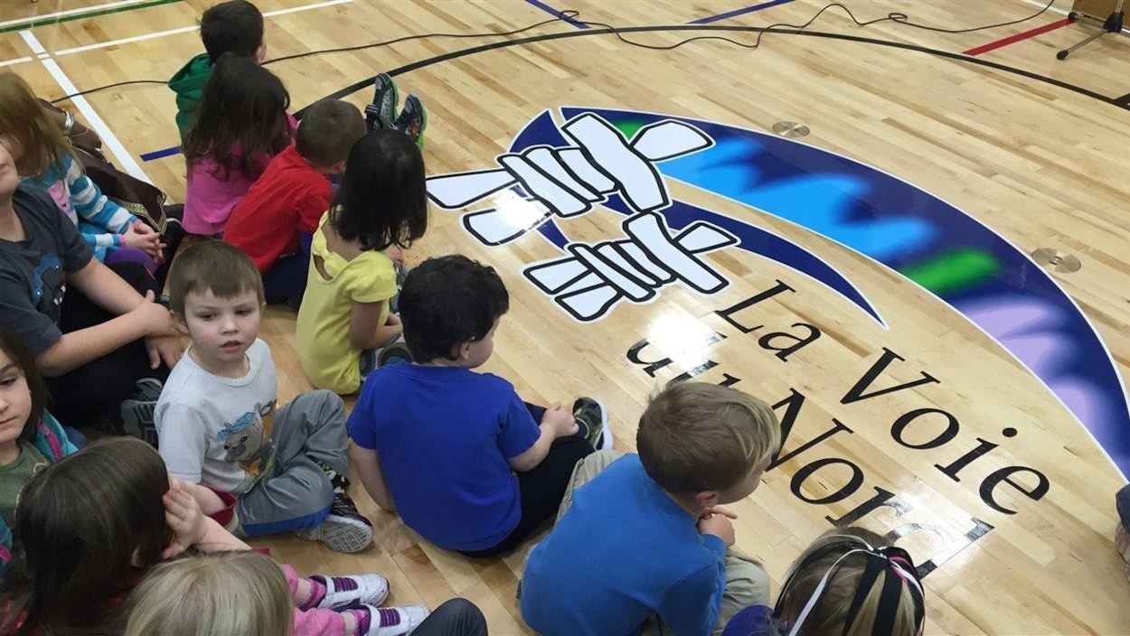 Des enfants assis par terre dans un gymnase. Sur le plancher est imprimé le logo de l'école, deux inukshuks avec des aurores boréales.