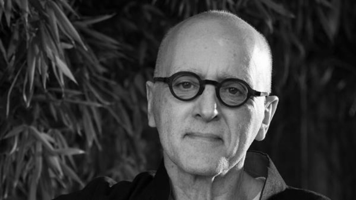 Sur une photo en noir et blanc, un homme, chauve, porte des lunettes rondes.