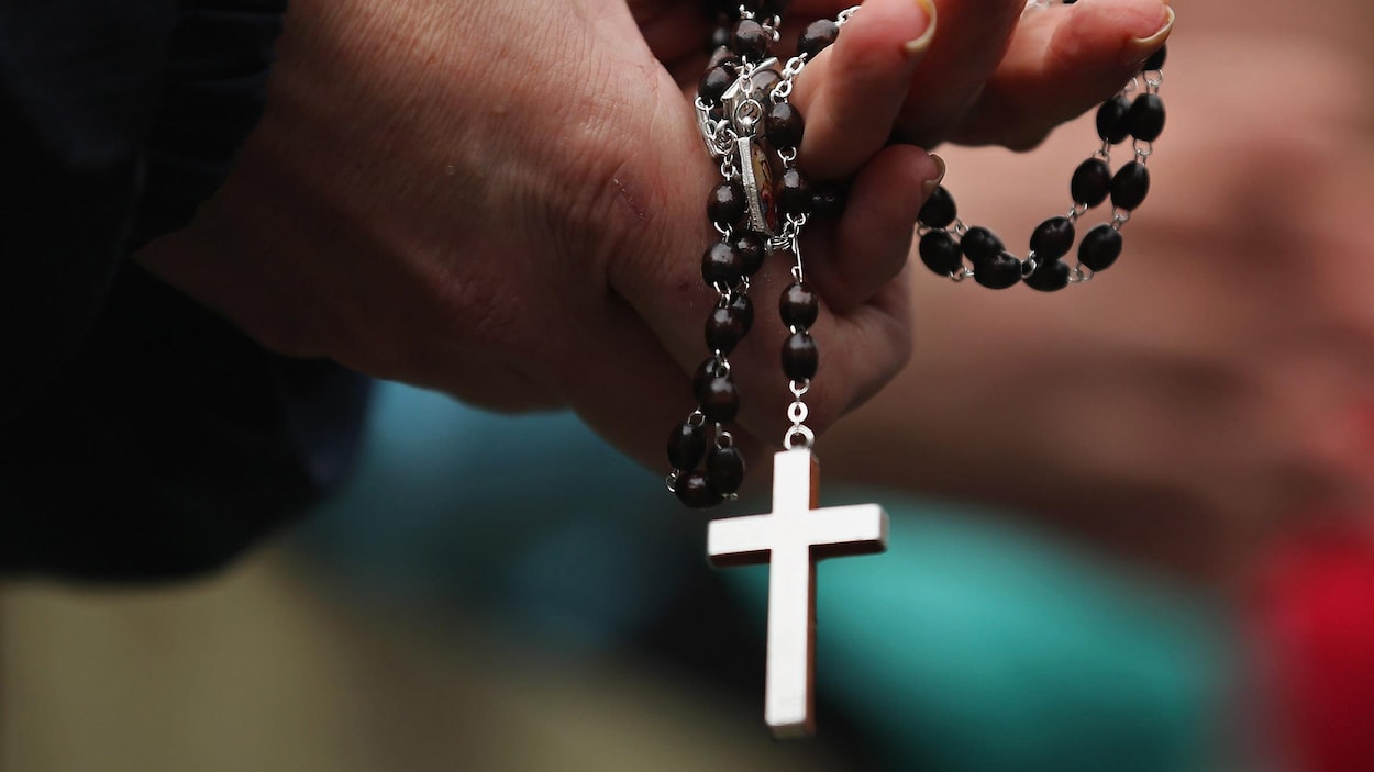 Les mains jointes, une paroissienne tient un chapelet avec, au bout, une croix bien visible au bout de la chaîne.