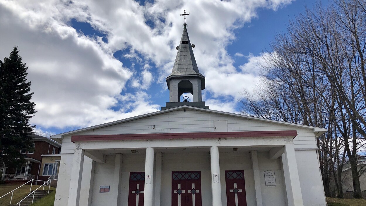 L'église est blanche et il y a des portes rouges. On peut voir au-dessus le clocher gris avec une croix.