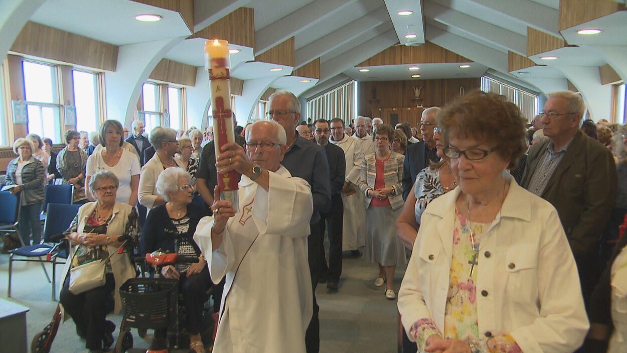 Un prêtre amène un cierge dans l'allée de l'église Saint-Jude. Des fidèles sont debout.