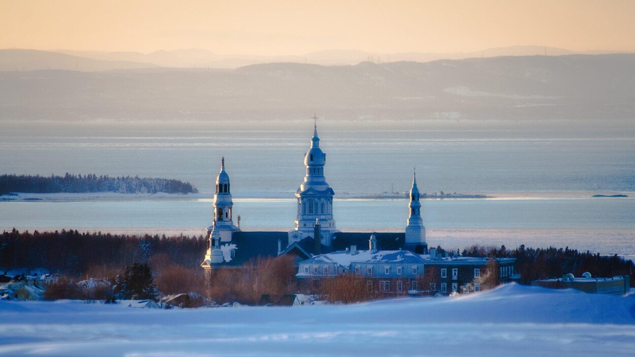 Point de vue sur une église qui se situe devant le fleuve Saint-Laurent, à Trois-Pistoles. On remarque les montagnes au loin qui trace une ligne d'horizon en hiver.
