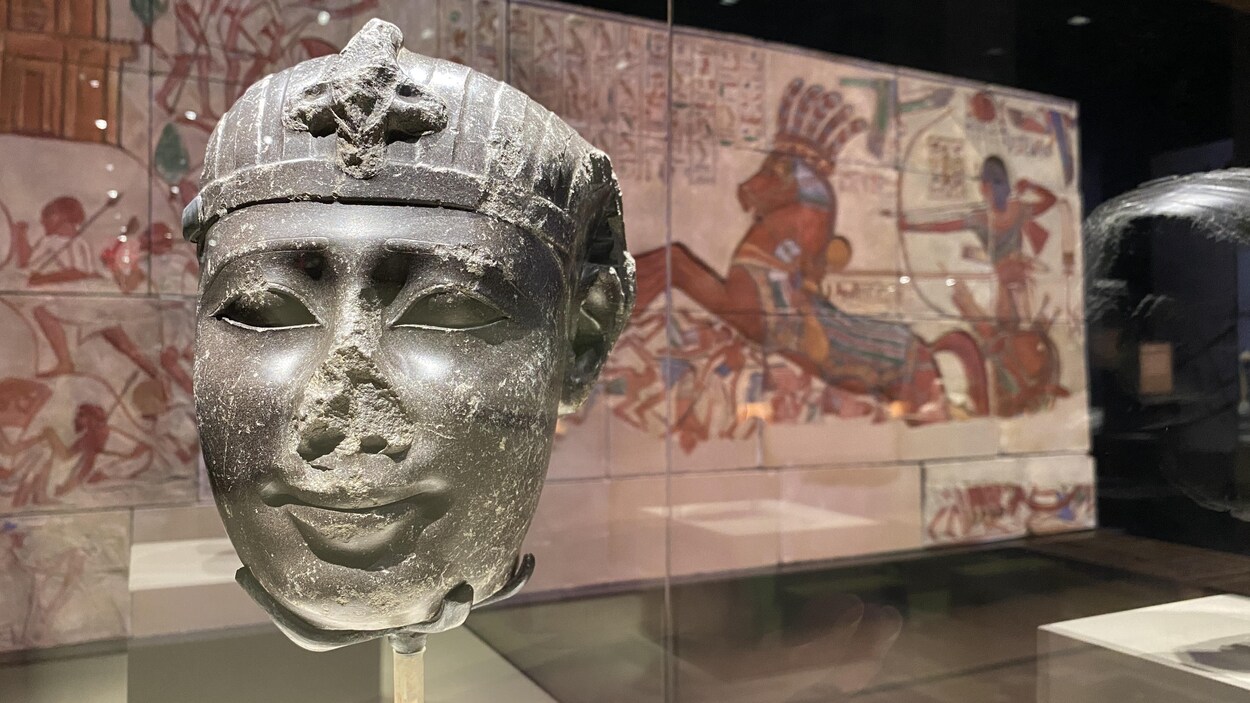Une tête de pharaon en pierre est installée devant une fresque égyptienne. 