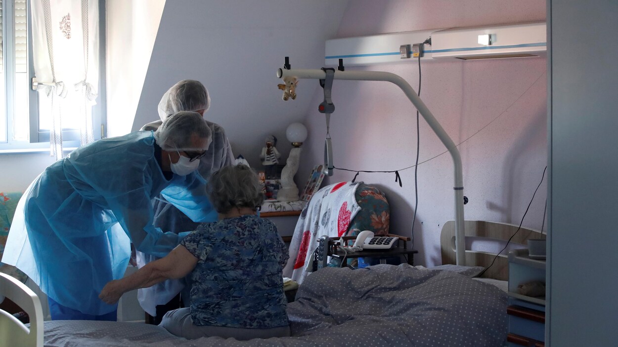 Deux personnes soignantes portant des vêtements protecteurs et un masque avec une aînée dans une chambre d'hôpital.