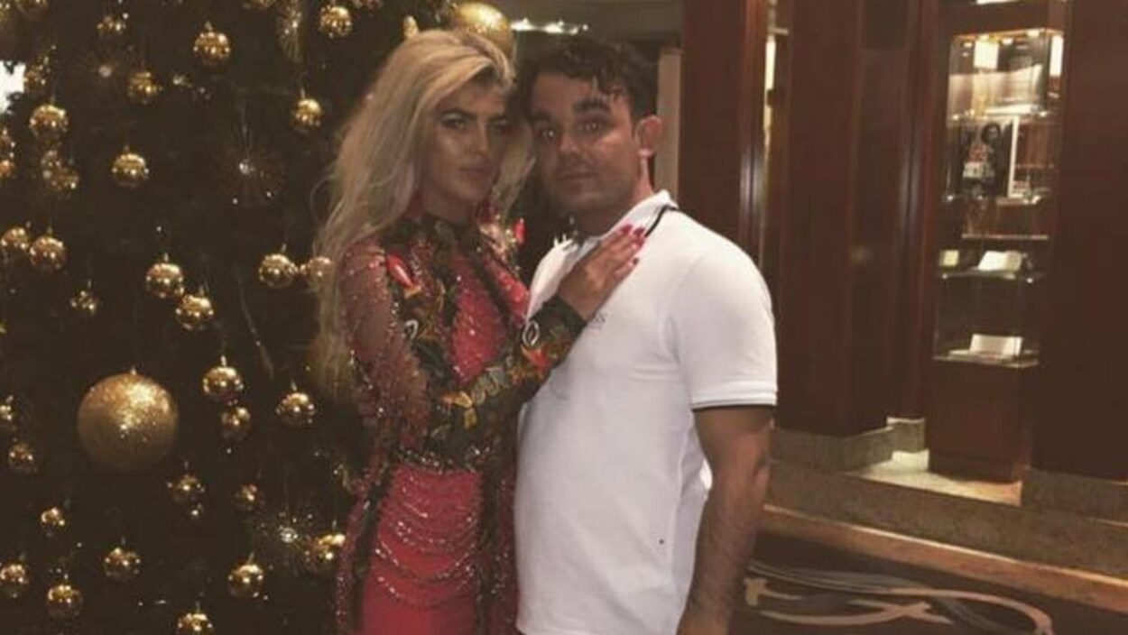 La jeune femme habillée d'une robe de soirée à perle, se tient debout aux côtés de son compagnon devant une sapin de Noël décoré.