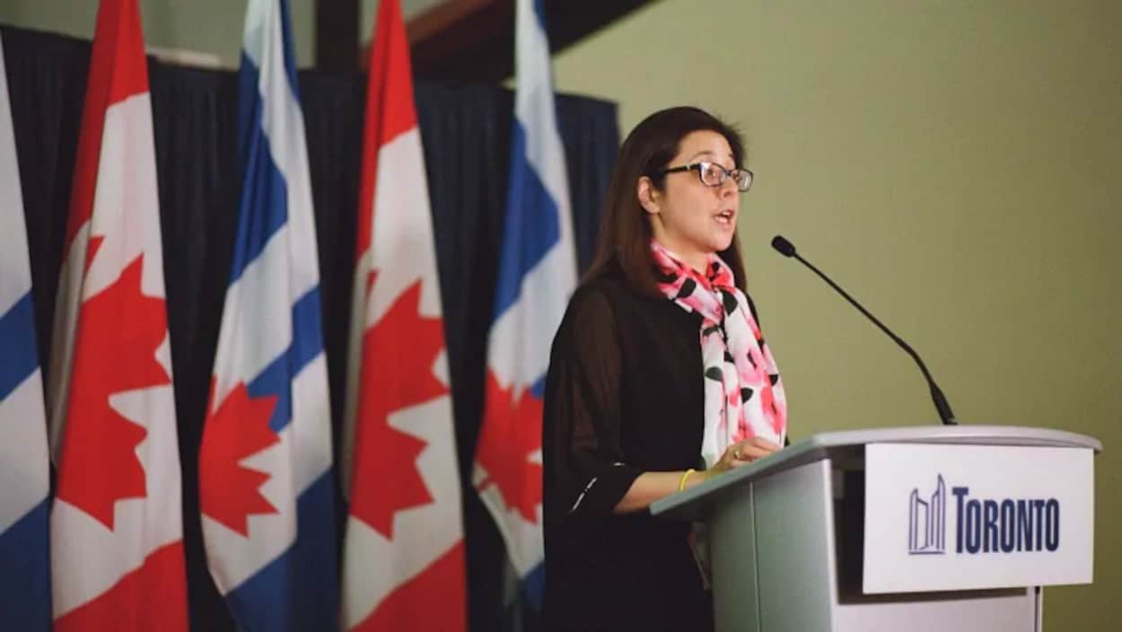 Elle parle à un lutrin devant des drapeaux du Canada et de Toronto.
